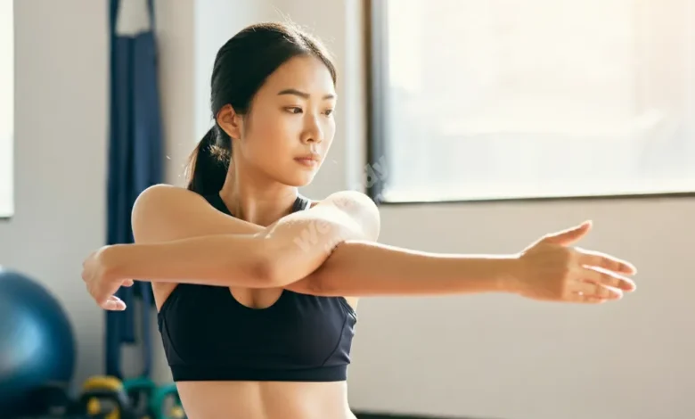 A person in athletic wear carefully stretching their arm, with a determined expression, in a bright, sunlit room with exercise equipment in the background